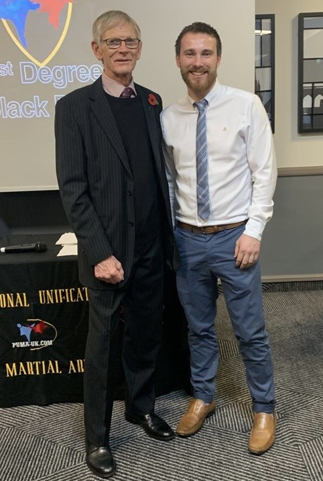 Sam Finch and Dennis Salt stood in front of a table with New Century Taekwon-Do on the table cloth and the logo on a presentation behind them